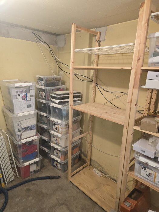 Hyllsystem i ett hobbyrum med plastlådor på hyllor, trähylla, gul vägg med synliga kablar och ett hörn av rummet syns.