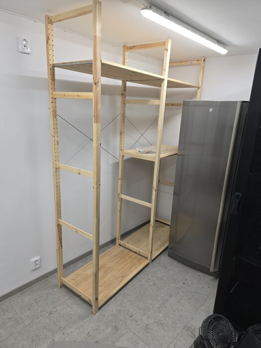 Trähylla av Ivar-typ står mot en vit vägg i ett rum med klinkergolv. Ett kylskåp syns till höger och en taklampa lyser ovanför.