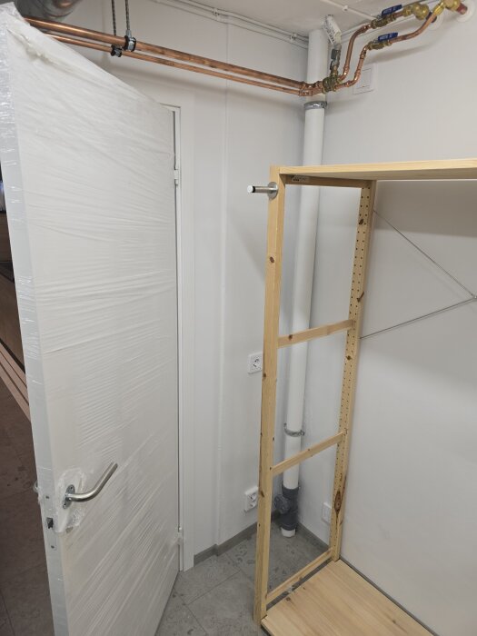 Öppnad dörr i hobbyrum med plastöverdrag, kopparrör på väggen och en delvis monterad trähylla under installation i hörnet.