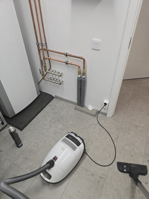 Dammsugare inkopplad i ett eluttag på en vit vägg med kopparrör, placerad på ett klinkergolv i ett maskinrum.
