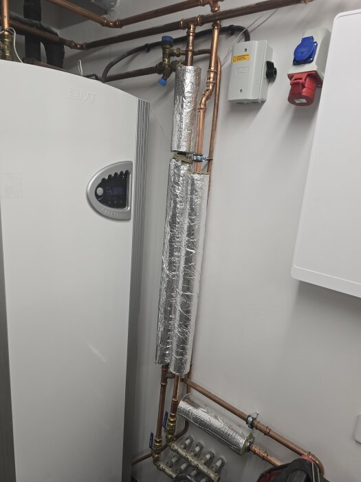 Maskinrum med rörsystem, inklusive kopparrör täckta med isoleringsmaterial. Elektriska uttag och en vit enhet synliga på väggen.