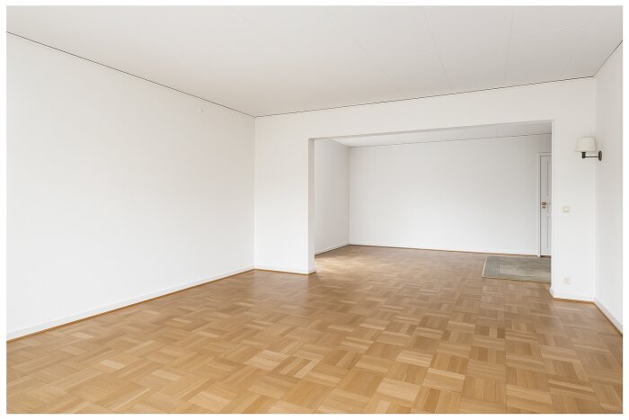 Tomt vardagsrum med parkettgolv och vita väggar, sett från glasdörr. Rummet har en öppen ingång till en annan sektion av huset.