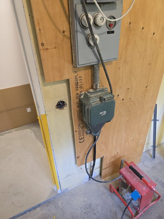 Elcentral monterad på plywoodskiva, med kablar anslutna, i renoverat utrymme. Golvborrning förekommer under installationen. En fläkt står på golvet.