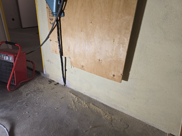 Plywoodskiva monterad på väggen med kabel, golv och en röd elektrisk apparat. Förberedelse för renovering.
