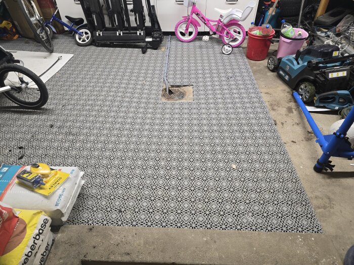 Bild av ett garagegolv med grått plastgolv och synlig golvbrunn. Runtom finns cyklar, verktyg och andra föremål.