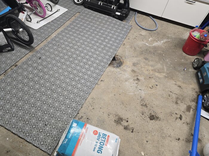 Betonggolv i garage med borttagen tröskel och synligt avloppssystem samt diverse föremål. En bit plastmatta saknas, golvbrunnen är synlig.