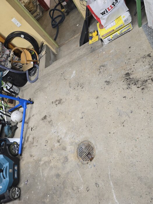 Golvbrunn av gjutjärn i ett garage med runtomkring liggande redskap och material samt en liten betongtrappa i bakgrunden.