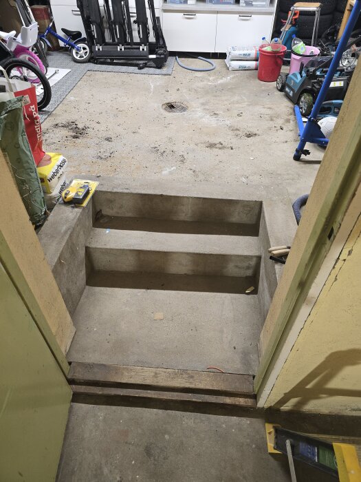 Betongtrappa i ett garage med borttagen tröskel och synligt avloppsrör. Verktyg och cykelhjul syns i bakgrunden.