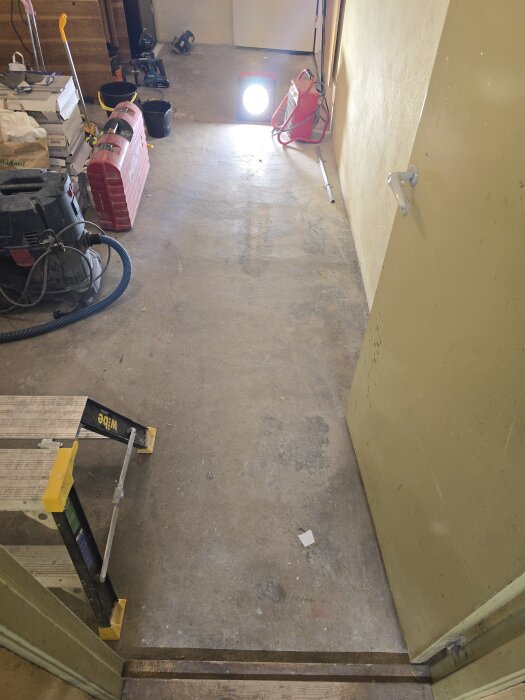 Garagegolv med verktyg och byggmaterial, en strålkastare lyser upp rummet, och en dörr är öppen, vilket avslöjar ett renoveringsprojekt pågående.