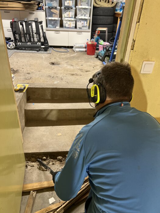 Person i blå tröja och hörselskydd arbetar med att ta bort en tröskel i ett garage, med verktyg i handen, omgiven av verktyg och föremål.