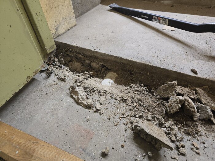Betonggolv med delvis grävt spår som visar ett grunt avloppsrör under en tröskel, omgiven av betongbitar och smuts. En kofot är synlig i bakgrunden.