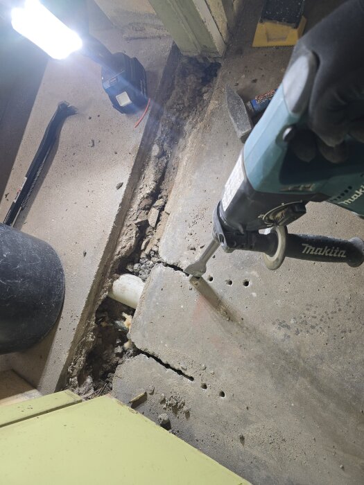 Man använder en hammare för att bryta upp ett betonggolv, synligt vitt avloppsrör under, omgivande byggmaterial och en arbetslampa.