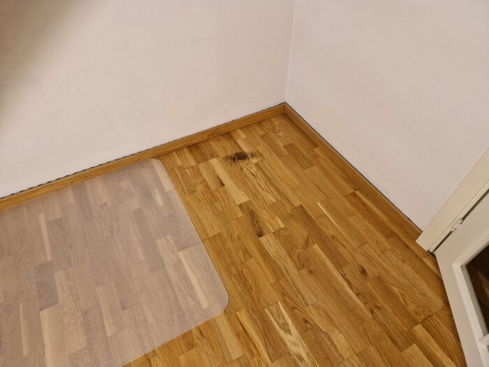 Trägolv med mörk fläck i hörnet, nära dörr och vägg; en plastmatta ligger delvis på golvet.