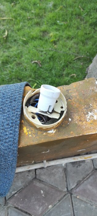 Ankelhög belysning på uteservering, med elkoppling inuti en rostig metallbox, omgiven av gräs och stenläggning.