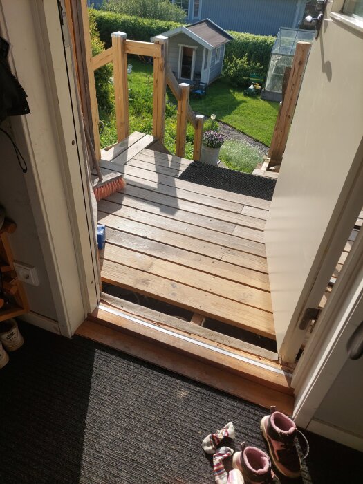 Öppen ytterdörr som leder ut till en träveranda med trappa. Soligt väder, gräsmatta och trädgårdsredskap syns i bakgrunden.