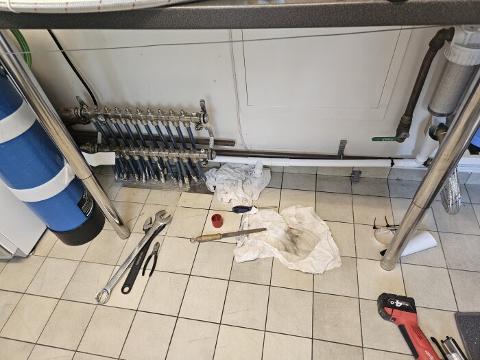 Rörinstallation under diskbänk med presskopplingar och verktyg som nycklar och tång på golvet, samt trasor.