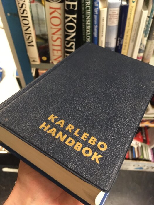 En hand håller en Karlebo handbok med mörkblå PVC-pärm framför en bokhylla med andra böcker i bakgrunden.