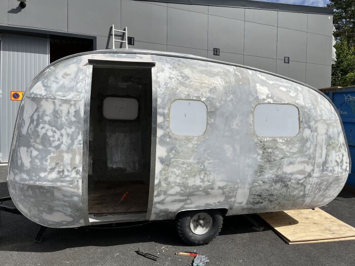 Renoverad husvagn med dörröppning, delvis slipad yta och två fönstergluggar. En stege lutad mot industribyggnad i bakgrunden.