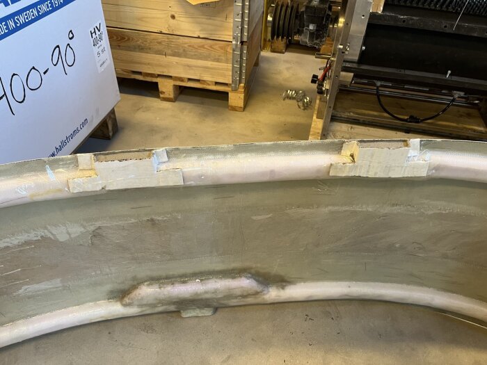 Plastad ficka på dörrbalk för dörrmontering, omgiven av tejpade aluminiumremsor, i verkstadsmiljö med trälåda i bakgrunden.