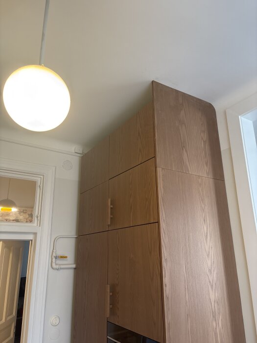 Högskåp i trä bredvid kyl och frys, del av köksrenovering. Ventilationsrör och dörr syns i bakgrunden.
