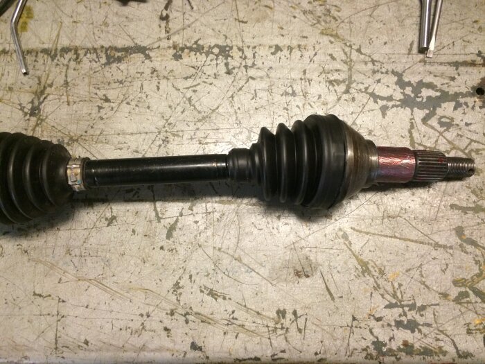 En drivaxel med stretch-damasker och metallfästen ligger på ett slitet arbetsbord, visar processen för reparation av ATV-delar.