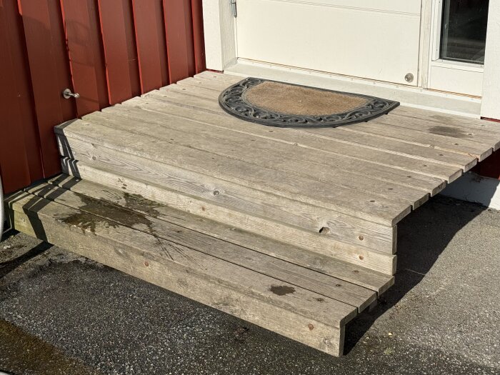Trätrappa framför dörr med röd fasad, planerad att ersättas med granit; en dörrmatta syns på översta steget.