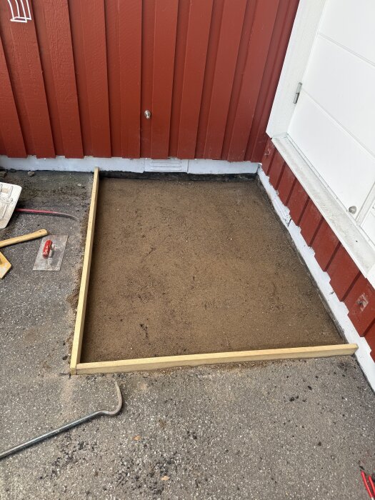 Form för granittrappa framför en röd träfasad och vit dörr, med borttagen asfalt och synlig sandyta. Verktyg syns på asfalten.