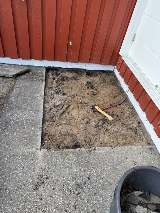 Grävd yta vid husvägg redo för granittrappa, verktyg och asfalt i bild.