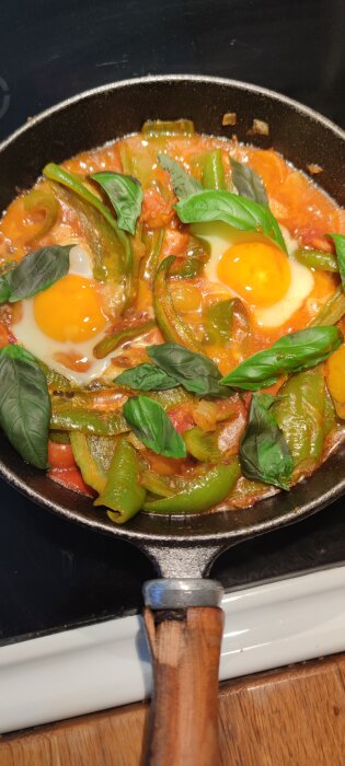 Stekpanna med shakshuka, innehållande två ägg, tomater och gröna paprikor, toppad med färska basilika blad.