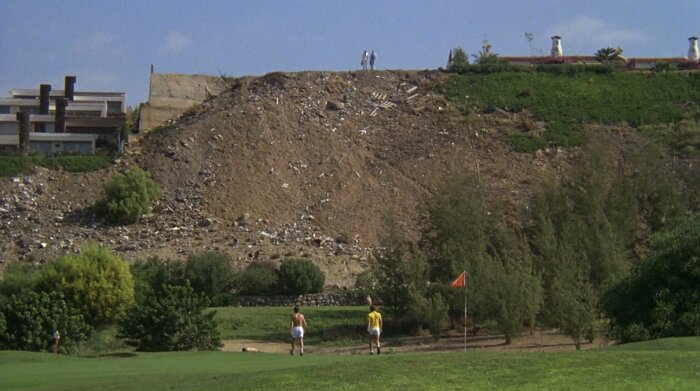 Två personer går på en golfbana framför en stor jordhög med skräp, med hus och träd i bakgrunden.