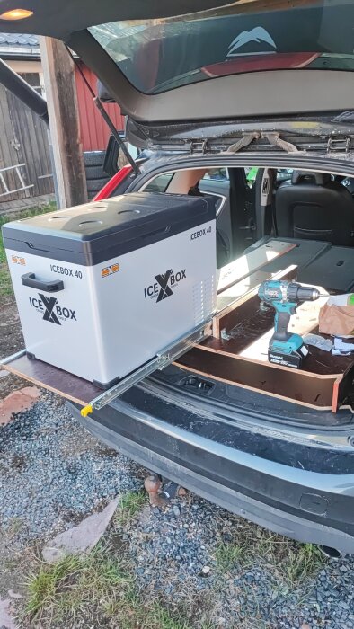 Bilens baklucka öppen med en kylbox och borrmaskin på utdragbar skiva.