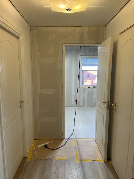 Renovering pågår i hall med öppna dörrar och väggar som förbereds för målning, en förlängningssladd ligger på golvet.