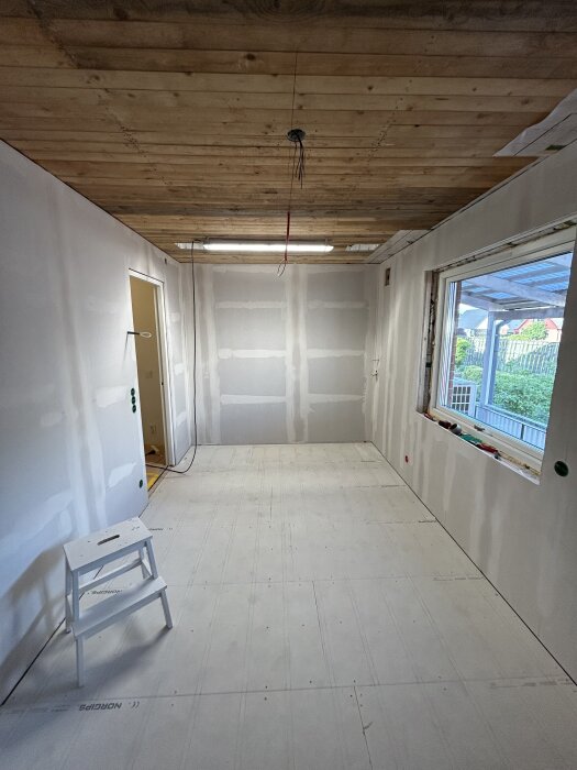 Rum under renovering med träpanel i taket, omålad gipsvägg och trappa vid dörren. Stort fönster med utsikt över en trädgård.