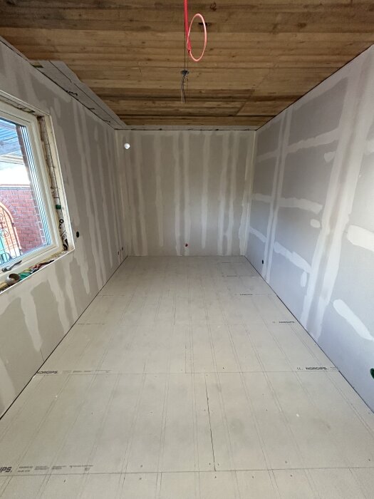 Nytt rum under renovering med gipsväggar och rått trätak. Tomt utrymme med ett fönster till vänster och hängande elkablar i taket.