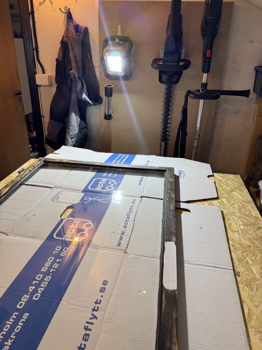 Målarbord med ett fönster som målas; bräda med vit färg, arbetsbelysning och upphängda trädgårdsverktyg på väggen.