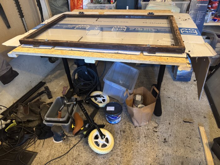 Ett målarbord med fönsterdelar i trä för målning, omgiven av färgburkar och verktyg i ett garage.