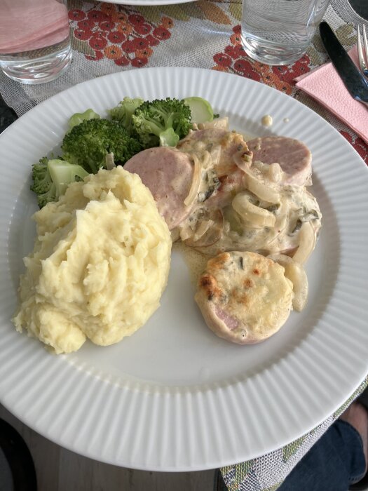 Tallrik med Bostonfalukorv i ugn, potatismos och broccoli, serverat på blommig duk.