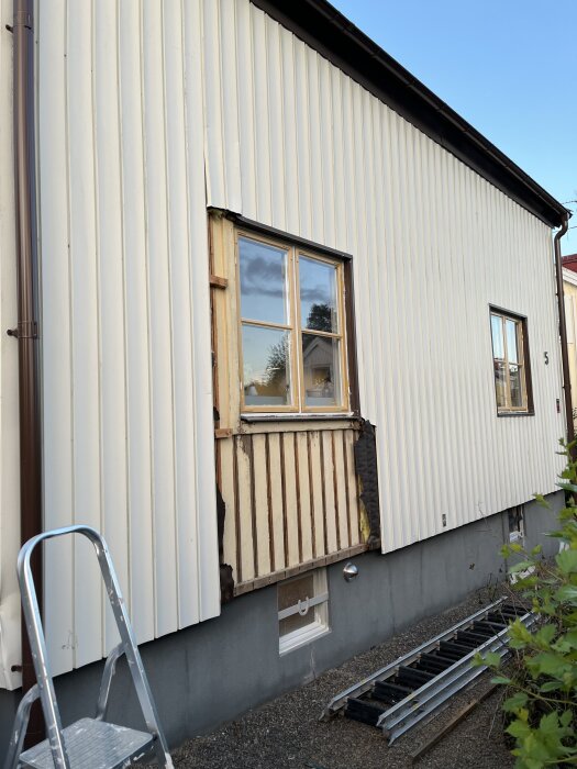 Vägg under renovering med borttagen panel runt fönstret. Stege placerad mot väggen, och en annan stege ligger på marken.
