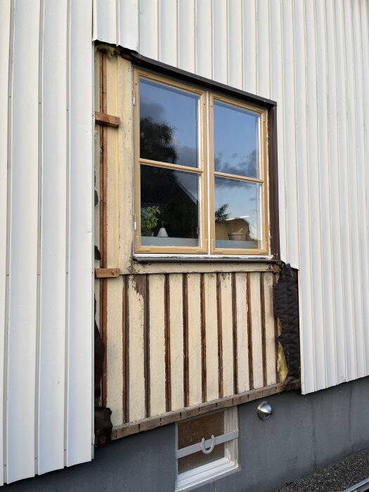 Renovering av husfasad med påbörjat arbete vid ett fönster. Träpanel och isolering synliga, mål  att få på läkten innan skyfall.