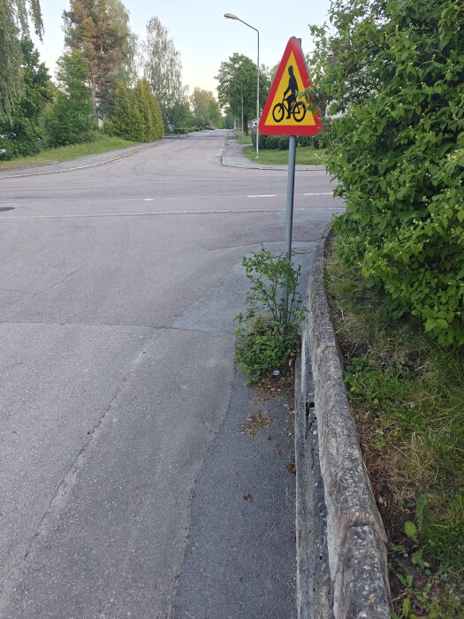 Gång- och cykelväg längsmed en gata med ett varningsmärke för cyklister, omgiven av grönska och en lågmur.