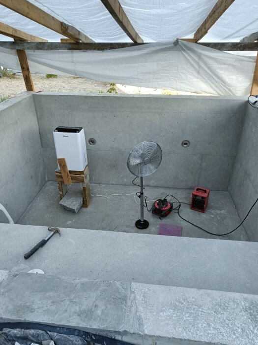Ofärdig pool med slipat betongskal, fläkt och avfuktare synliga. Provisorisk presenning över öppningen, olika verktyg på kanten.
