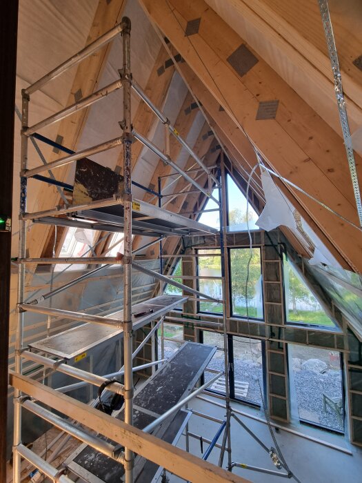 Byggställning med plattformar och gavlar inuti ett trähus under konstruktion, med utsikt genom stora fönster mot naturen utanför.