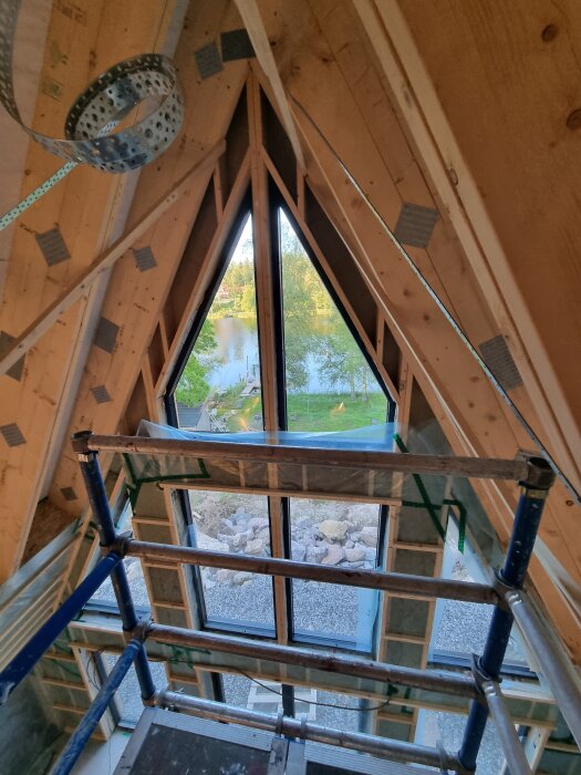 Byggnadsställning uppställd inomhus framför ett stort fönster med utsikt över en sjö och omgivande skog.