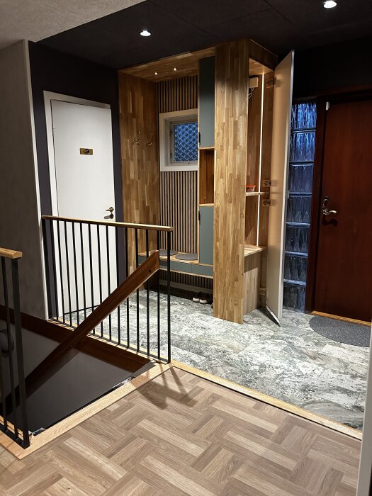 Trappa med räcke 100 cm högt, modernt trä- och metallutformning, intill en hall med klinkergolv, garderob och dörr.