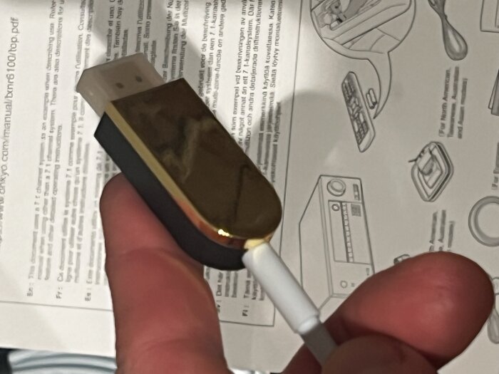 En person håller i en HDMI-kabel med en guldfärgad kontakt, framför en teknisk manual som visar bilder av elektronisk utrustning.