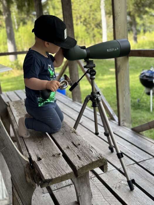 Barn med keps tittar genom en kikare på ett träbord utomhus vid en sommarstuga.