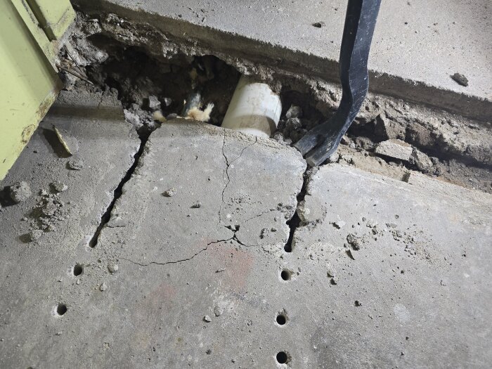 Bilad betongplatta med synligt avloppsrör; spruckna ytor och en kofot vid kanten, grönt skåp till vänster.