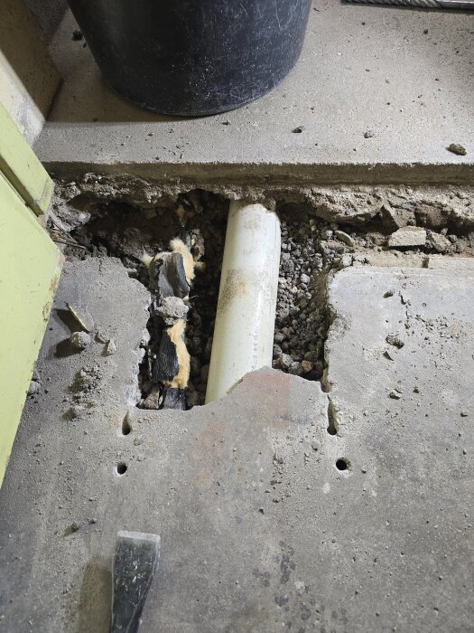 Avslöjad avloppsledning under ett betonggolv, omgiven av sand och grus. En liten kofot är synlig på kanten av det brutna betonglagret.