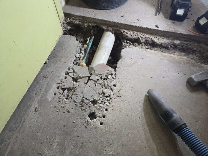 Betonggolv med uppbrutet område och synligt avloppsrör, omgiven av verktyg som en hammare och en dammsugarslang.