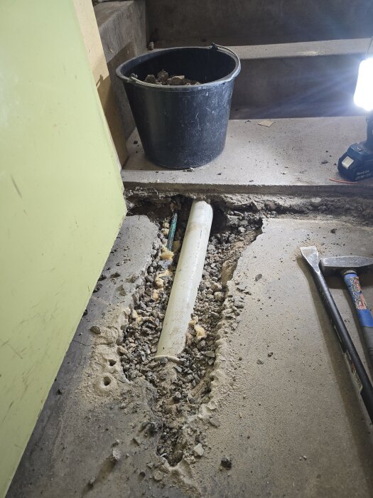 Avtäckt avloppsrör i betonggolv vid renovering, omgiven av grus och verktyg, med en svart hink och belysning i bakgrunden.